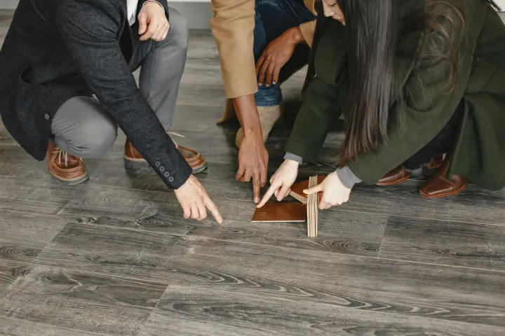Three people kneeling on a wooden floor, comparing flooring samples